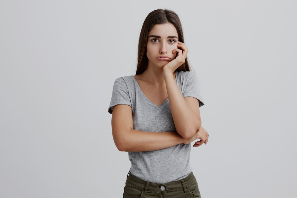 A woman with long hair wearing a gray shirt wondering with her hands on her chin