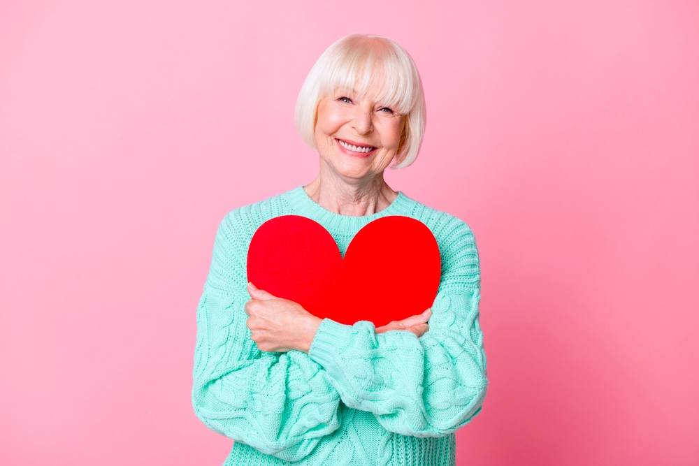 A woman with green sweatshirt hugging a heart shape in pink background.